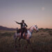Arthur Morgan on a dapple gray Thoroughbred in a meadow. He points a revolver at someone out of frame. Behind him, the moon is beginning to appear as the sun goes down.